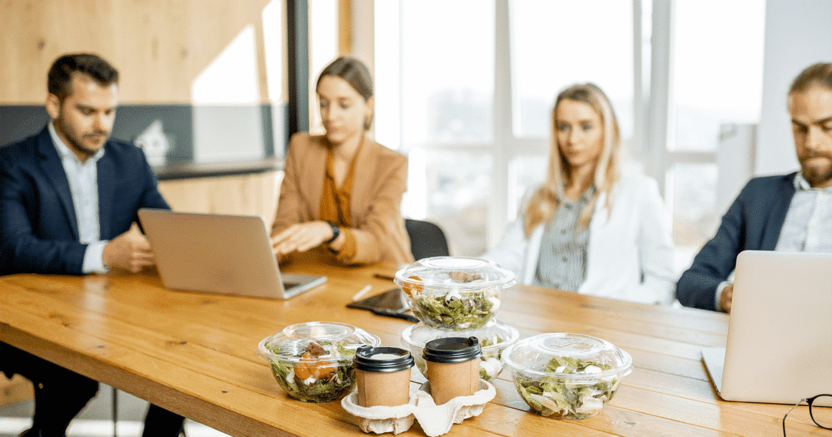 Kantoortafel met salades, drankjes en vier mensen op de achtergrond